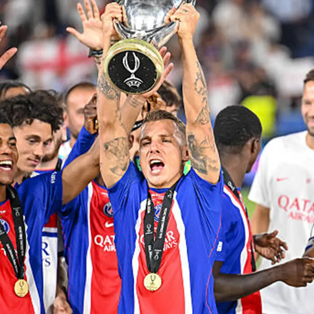 A football player in a blue, red, and white PSG jersey lifts a large silver trophy above his head in a celebratory moment, surrounded by teammates wearing Qatar Airways jerseys.
