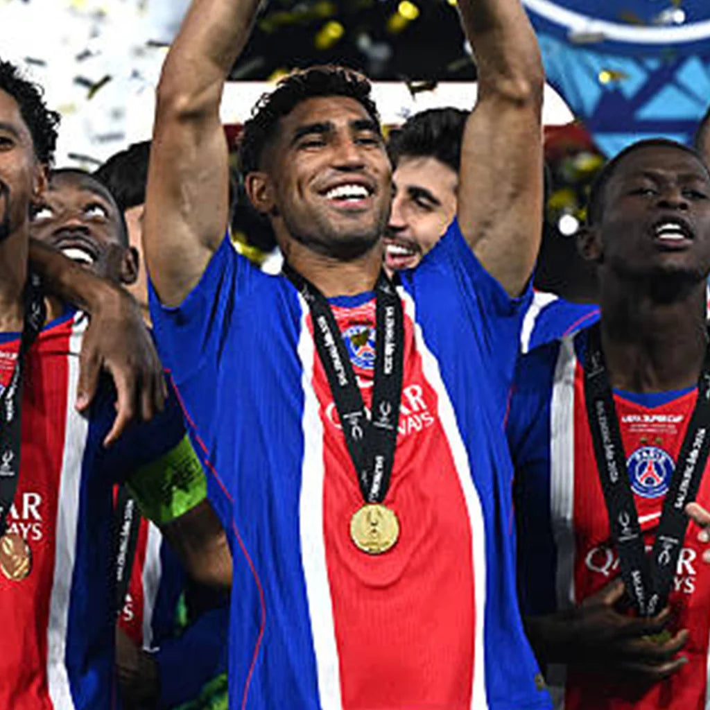 A soccer player in a blue, red, and white striped jersey celebrates with teammates on a podium, wearing a gold medal around his neck. The Paris Saint-Germain logo is visible on the jersey and medals.