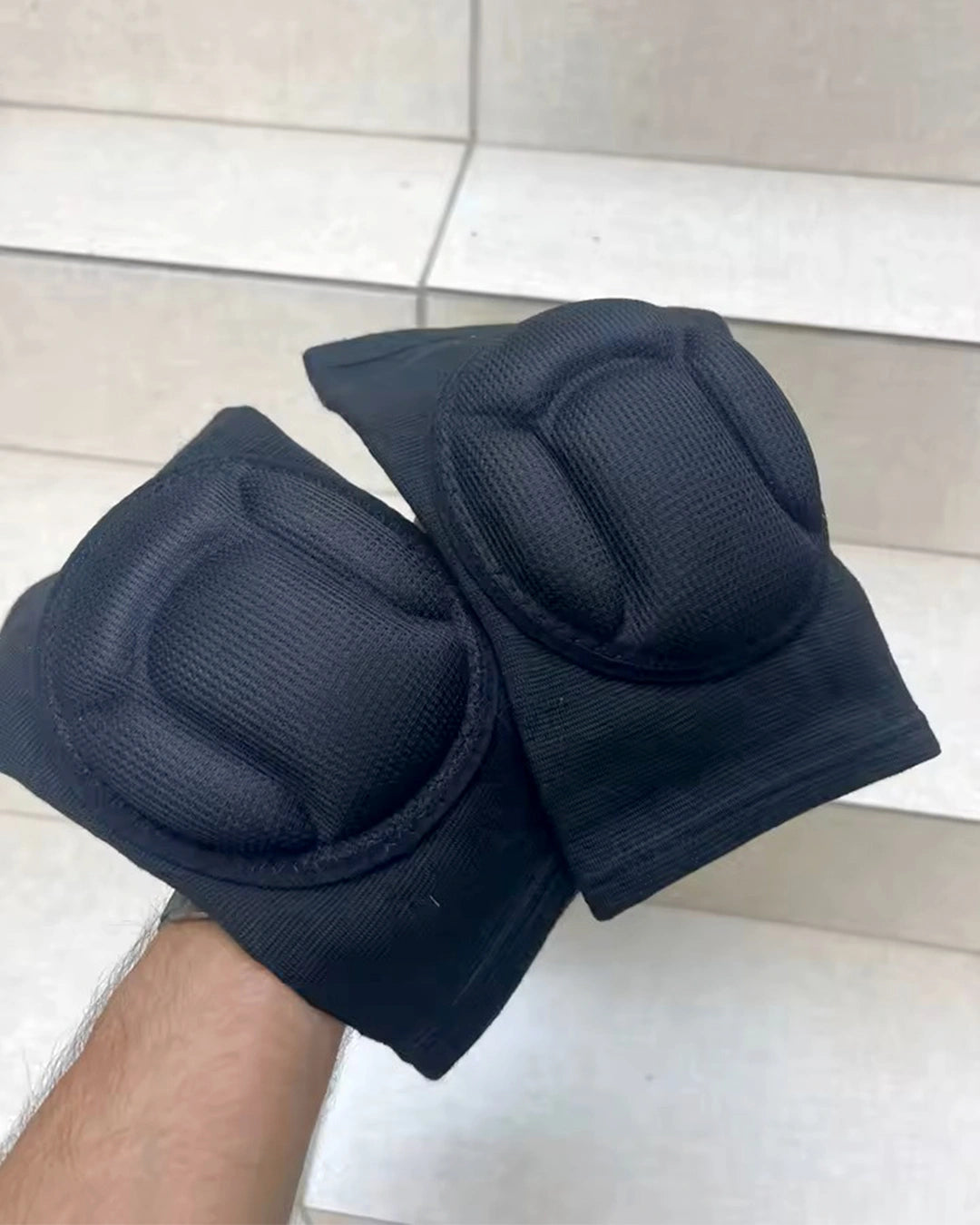 Black knee pads held by a person against a tiled wall background