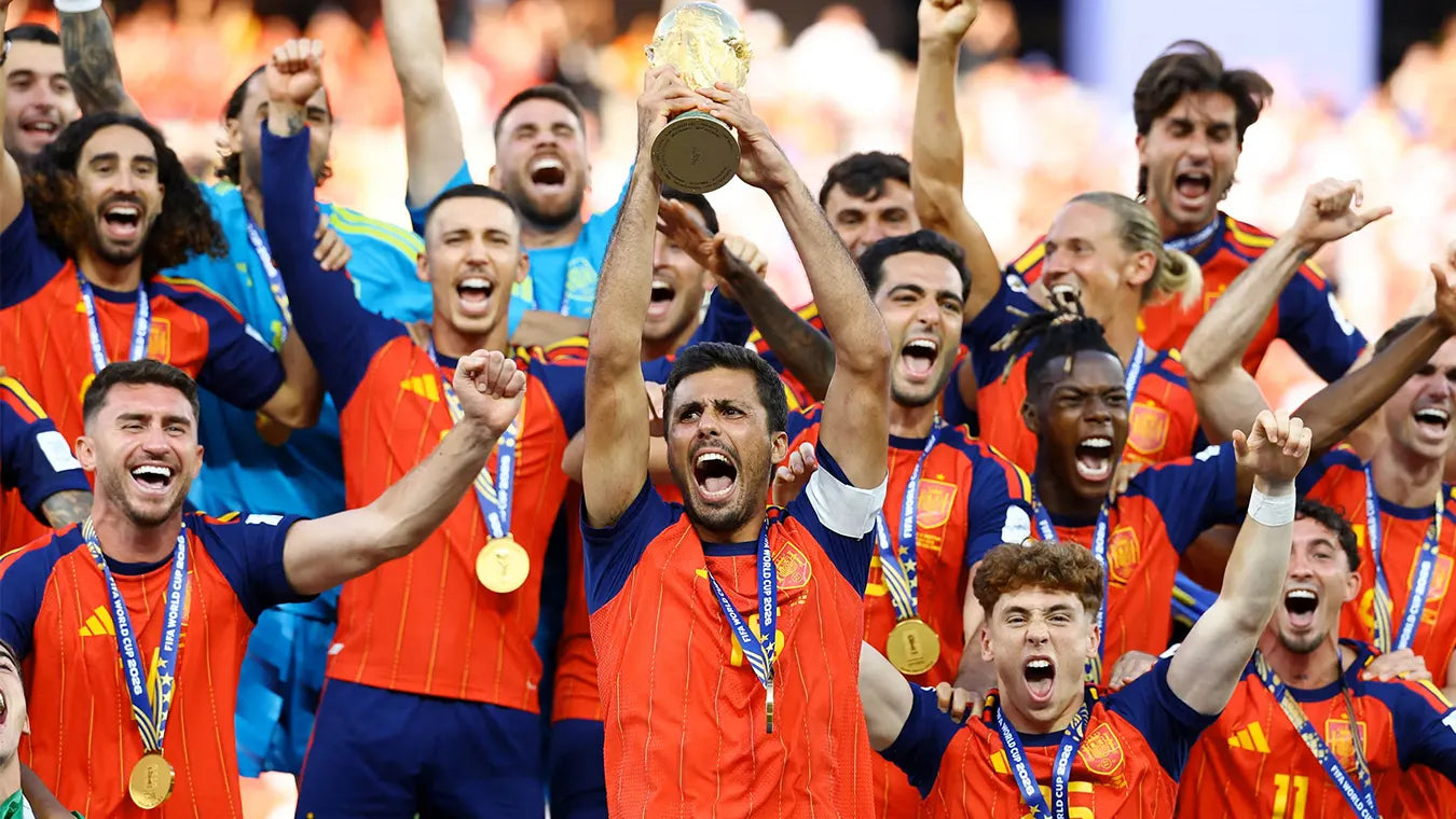 A group of soccer players in Spain national team orange and navy jerseys celebrate with raised arms and gold medals, holding a trophy in a stadium setting.